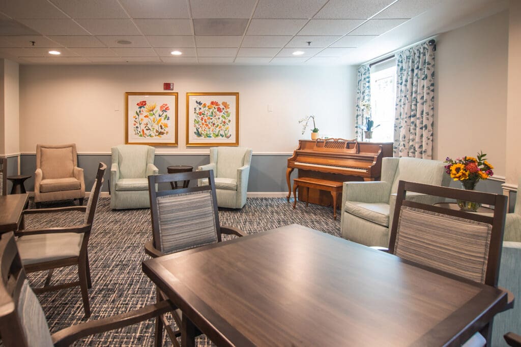 An image a communal living room at Charter Senior Living of Towson, featuring tables, chairs, and a brown piano.